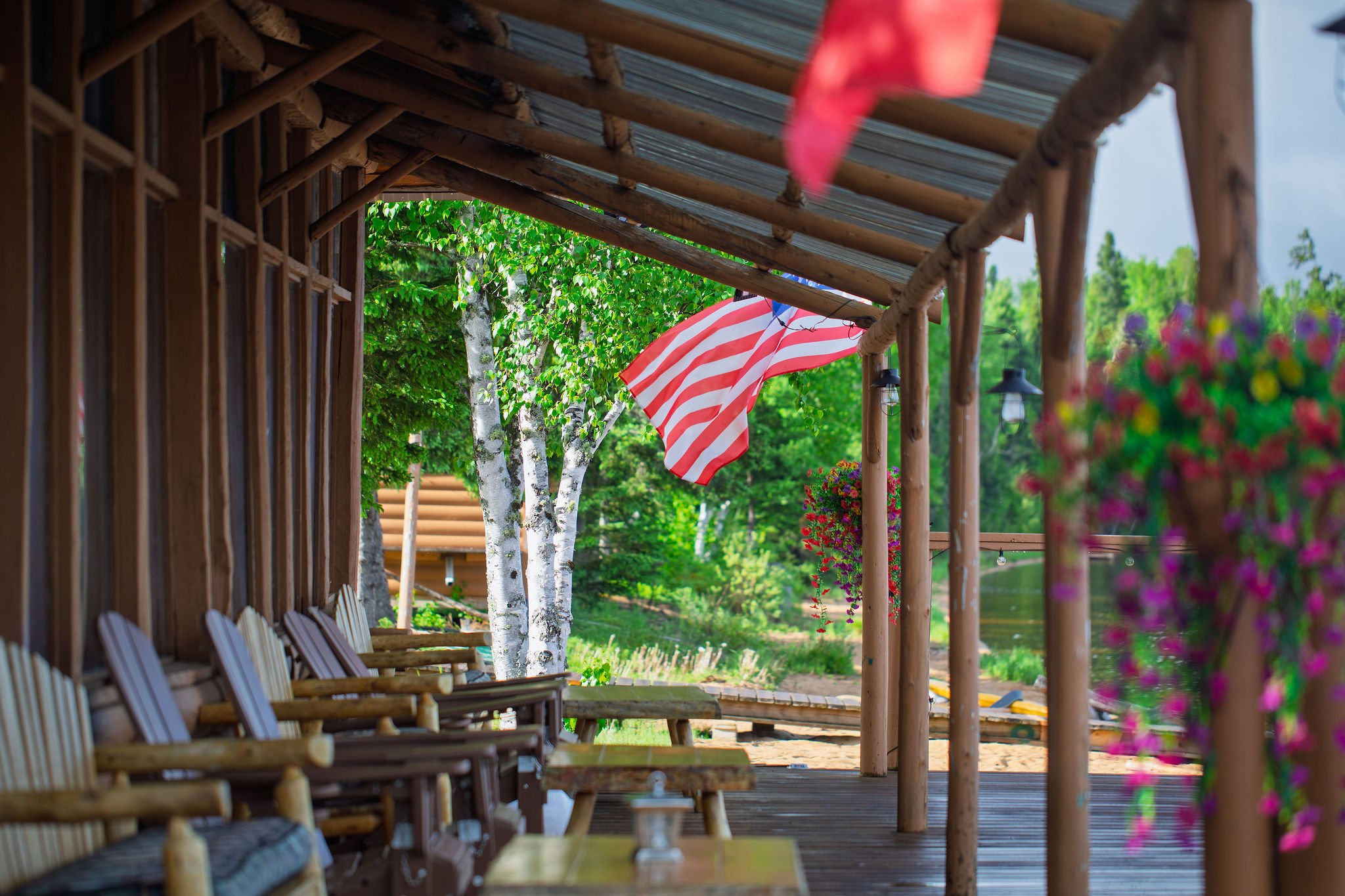 The wooden porch of Cat Island Lodge in Ontario, Canada, with row of wooden chairs along the side, led by a wooden railing and posts, with a view of green trees, a colorful striped umbrella, and hanging lanterns outdoors.