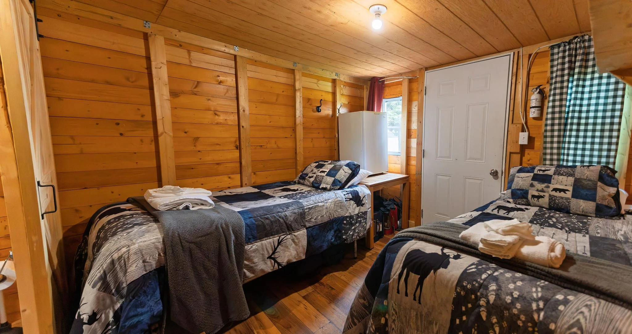 A rustic bedroom with wooden walls and ceiling, two twin beds with bear-themed quilted bedding, a small window with a red curtain, a white door, and a fire extinguisher mounted on the wall.