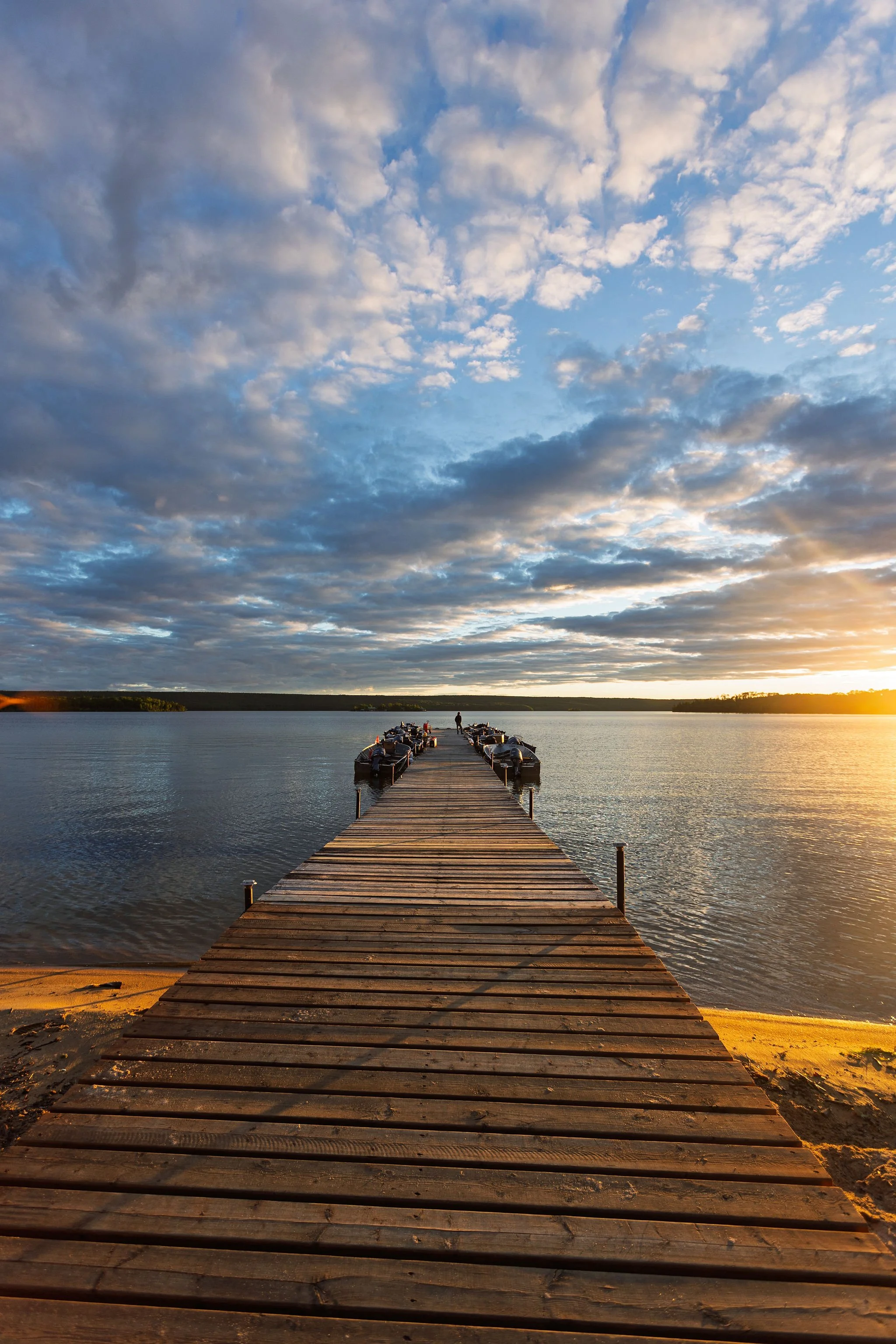 Wooden dock extending into a calm body of water during sunset, with boats docked on either side and a person standing at the end of the dock, under a partly cloudy sky.