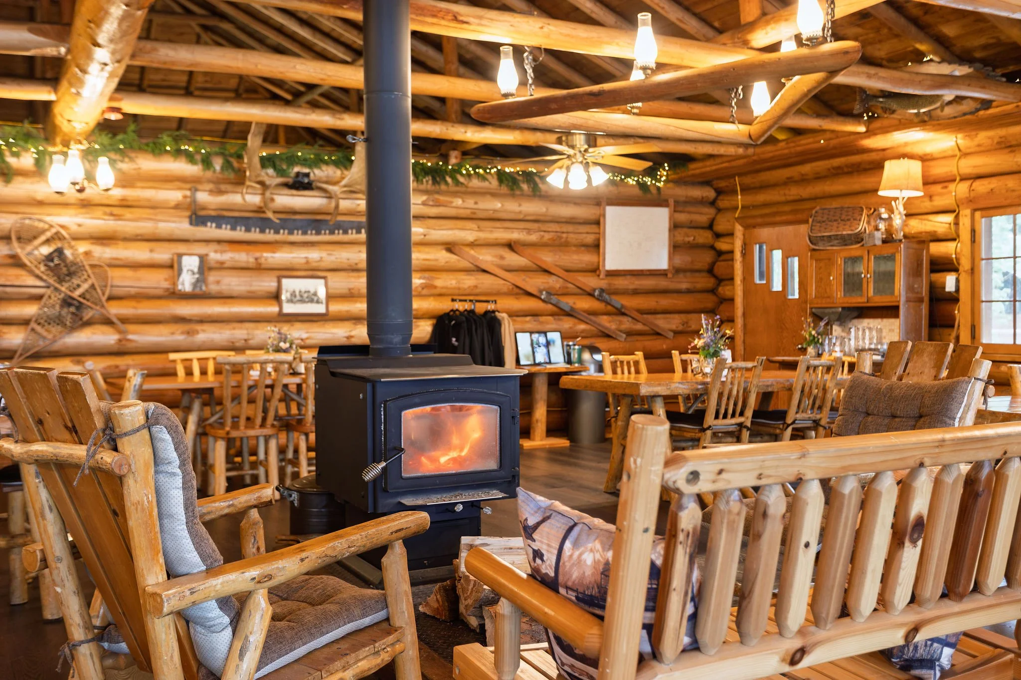 Interior of a rustic log cabin with wooden furniture, a central wood stove, and decorative lighting including lanterns and string lights. There are framed pictures and greenery along the logs, creating a cozy atmosphere.