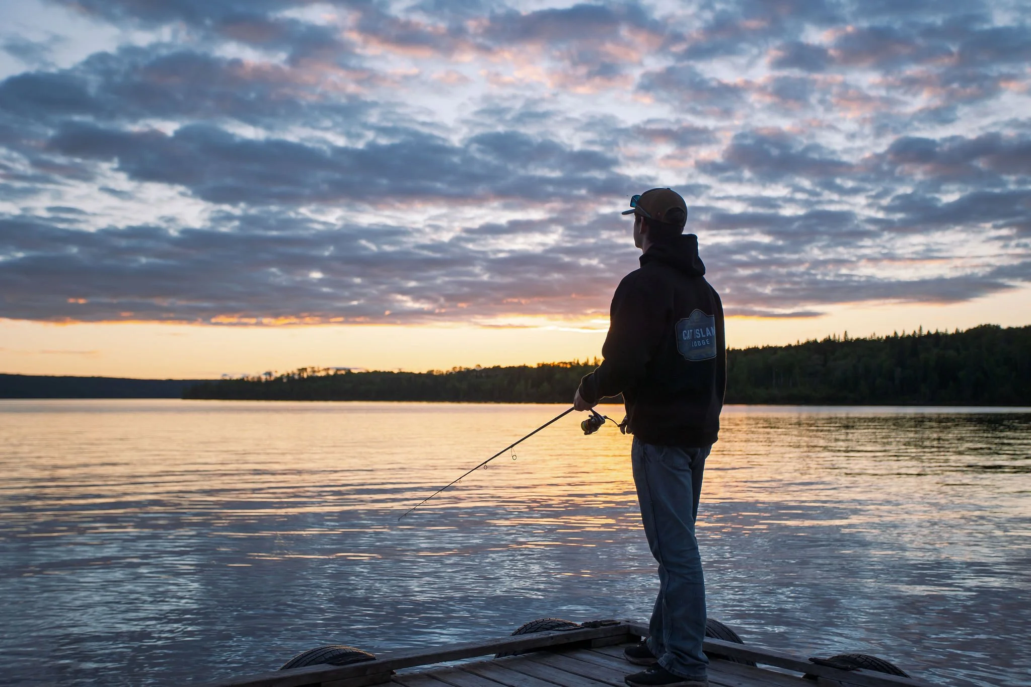 A person standing on a dock at sunset, fishing in a lake. The person is wearing a hoodie, cap, and sunglasses, with a cloudy sky overhead.