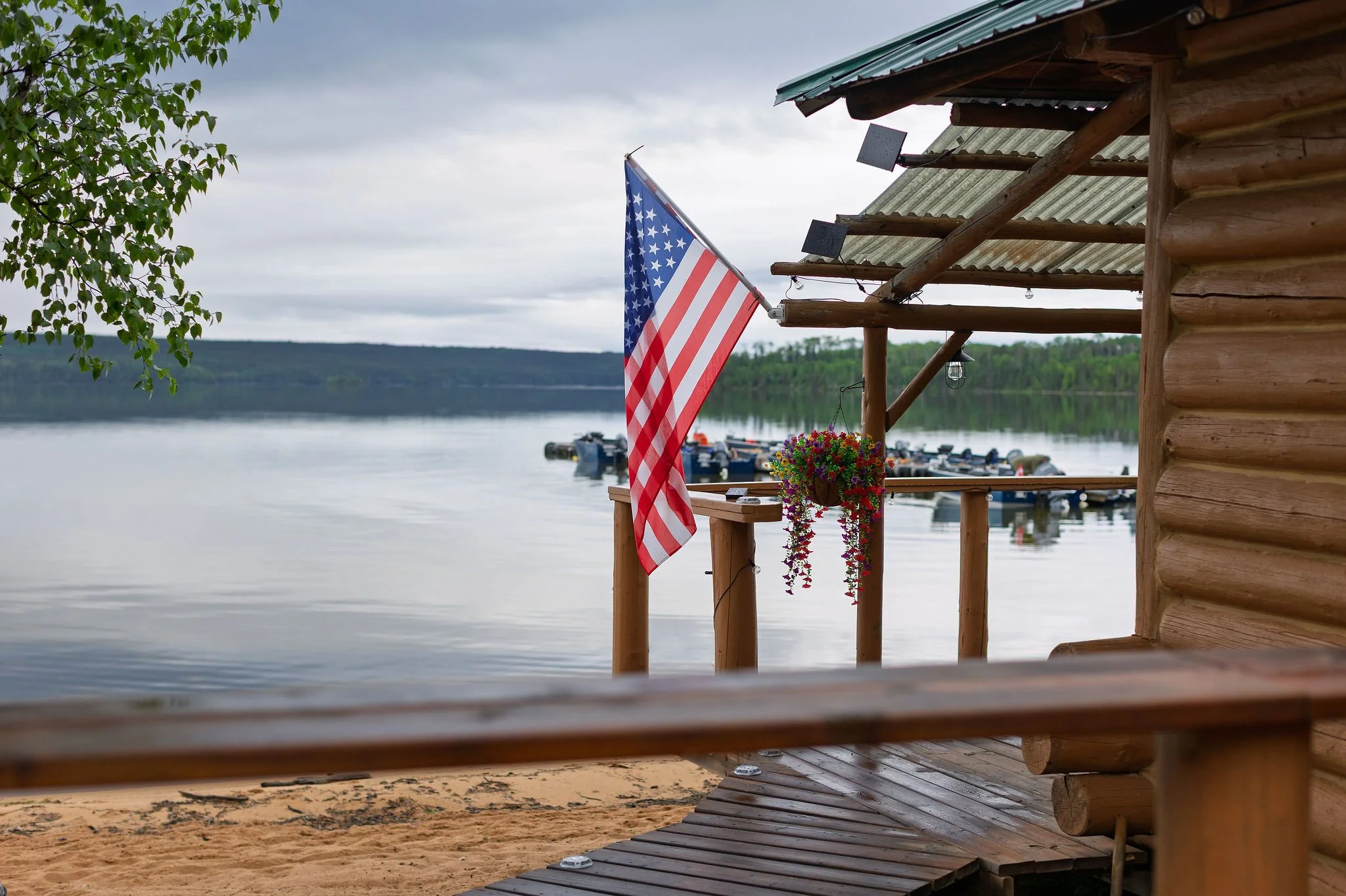 A wooden porch with an American flag hanging, overlooking a serene lake with boats and distant hills under a cloudy sky.
