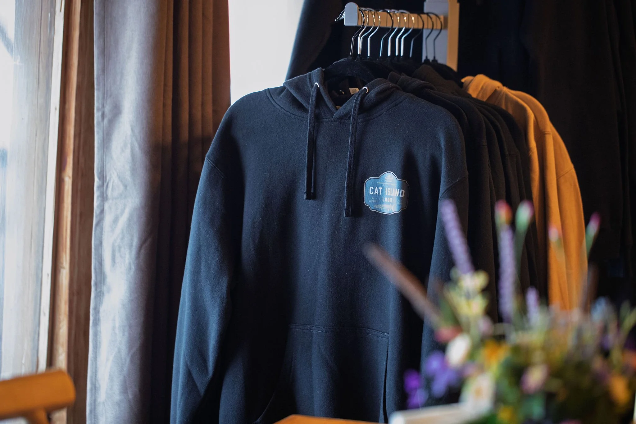 Hoodies in black and yellow hanging on a rack in a store next to a window with curtains, with out-of-focus plants in the foreground.