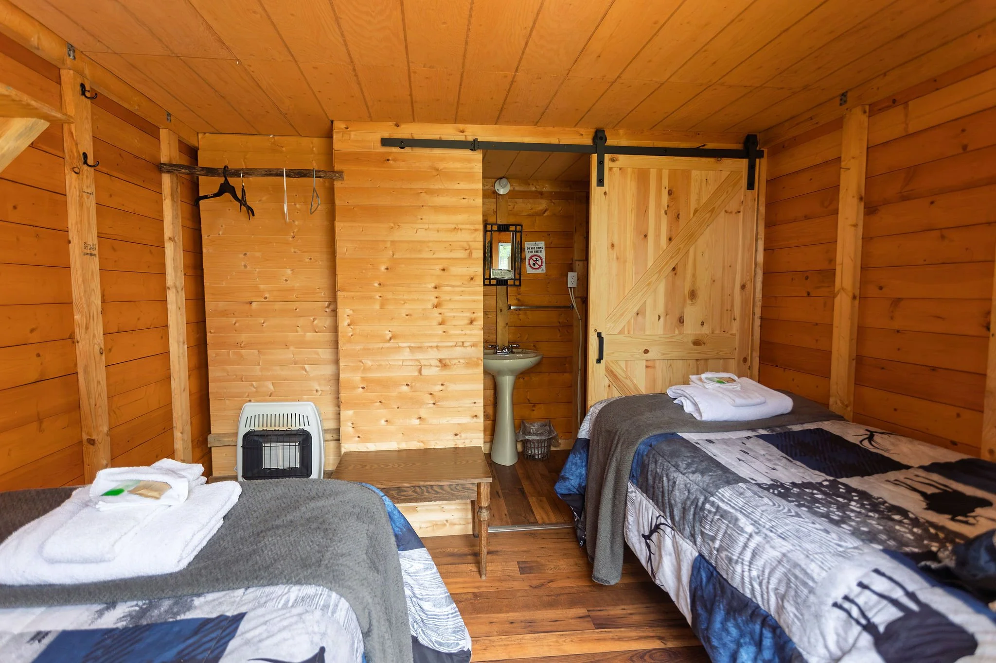 A cozy rustic bedroom with wood-paneled walls and ceiling, featuring two beds with animal-themed bedspreads, one with folded towels, a small heater, a chair, a small sink, a mirror, and a sliding barn-style wooden door.