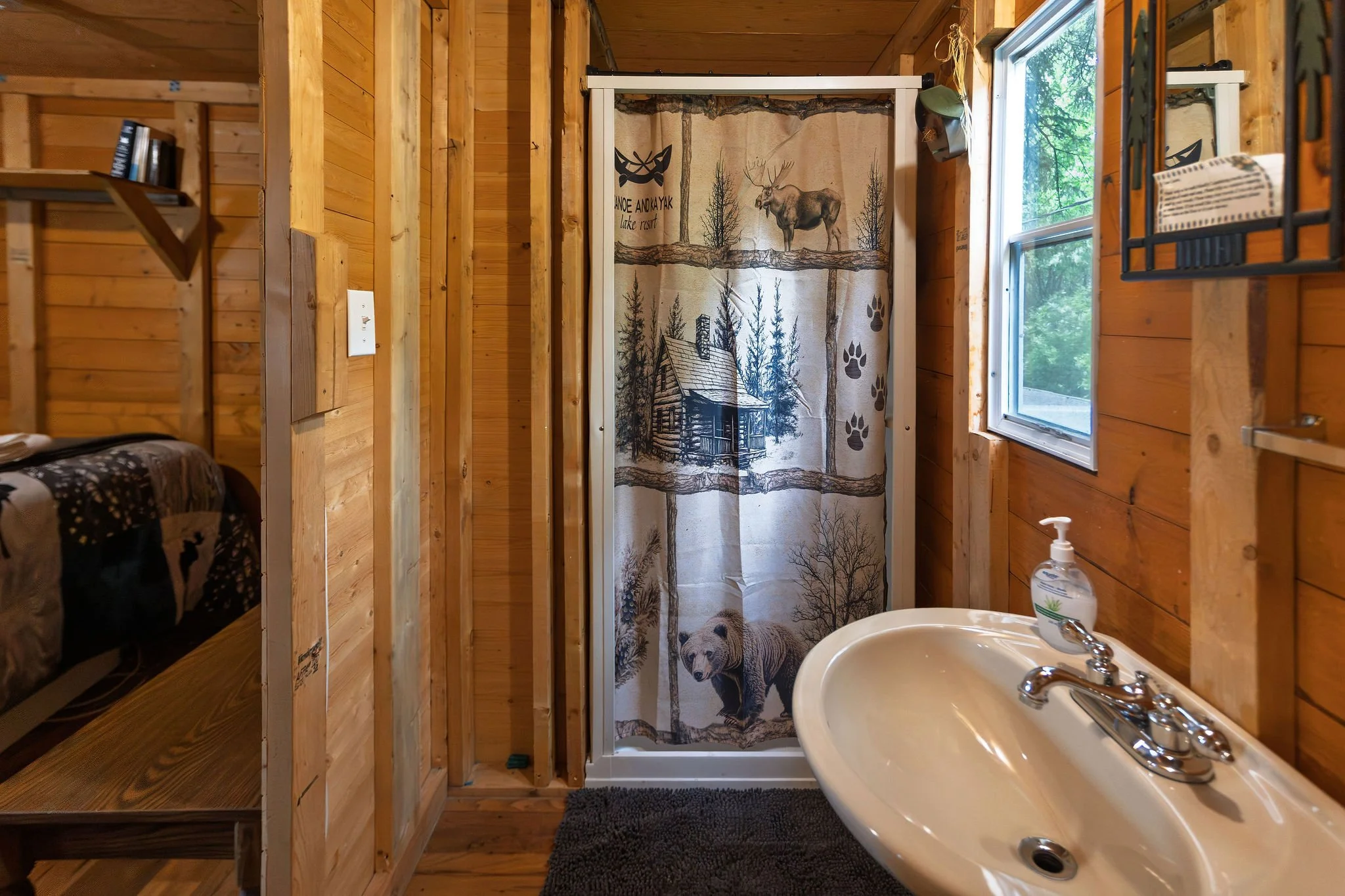 Interior of a rustic wooden cabin bathroom with a small sink, a soap dispenser, a window with trees outside, partially visible bed, and a shower with a curtain featuring outdoor forest and wildlife scene.