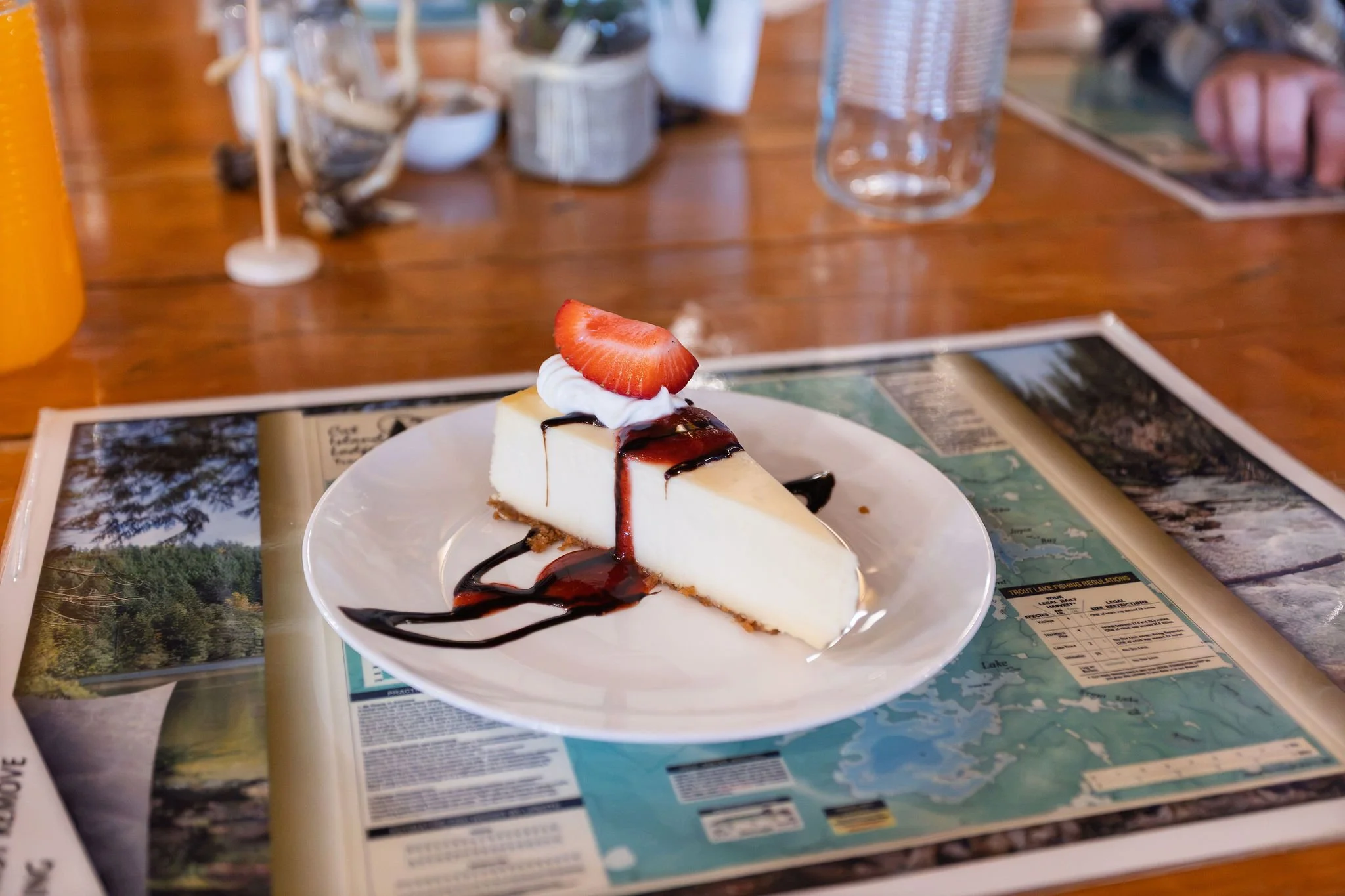 A slice of cheesecake with a strawberry on top, drizzled with chocolate syrup, on a white plate. The plate is on a table covered with travel maps and magazines, with a blurred background of a wooden table, glassware, and decorative items.
