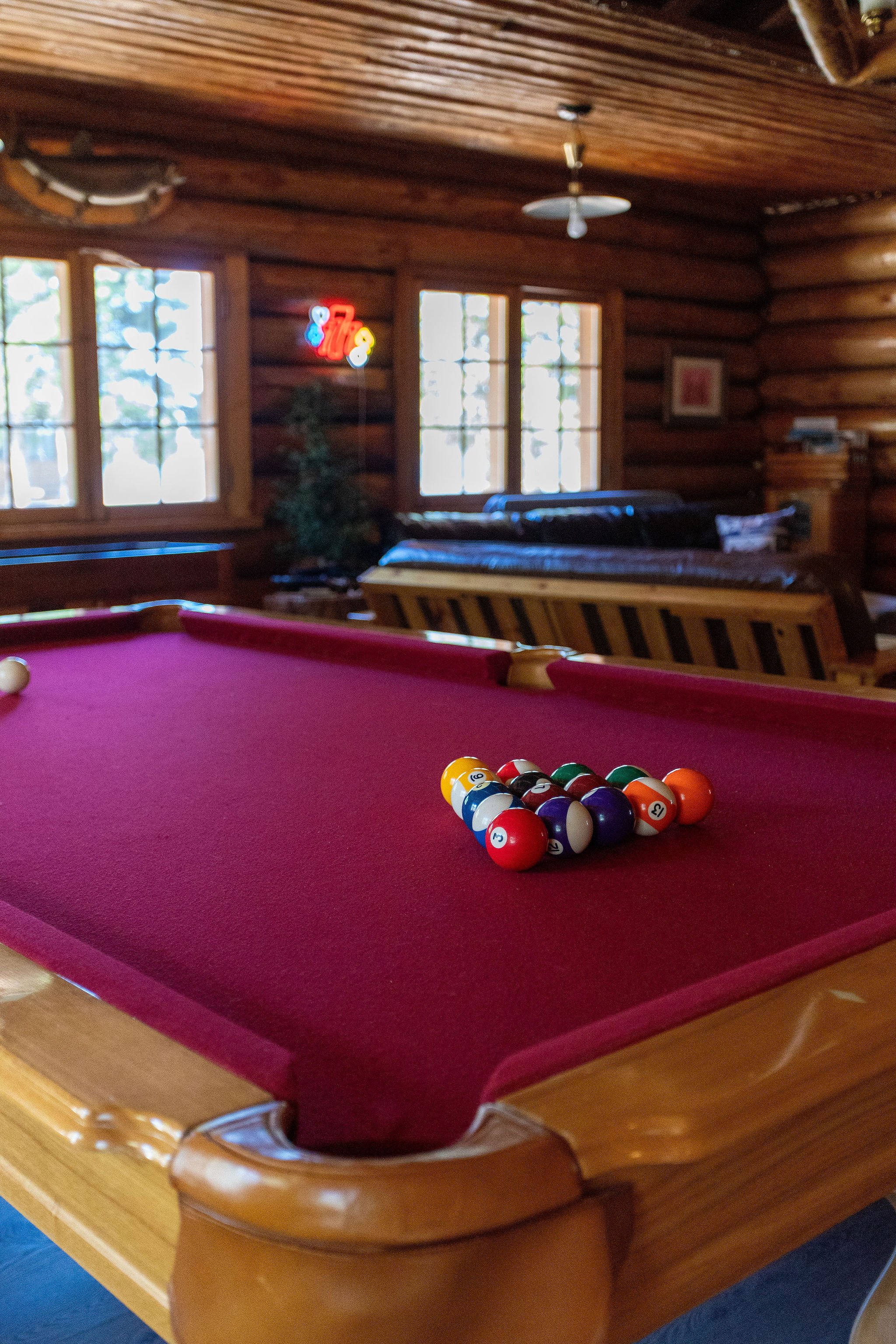 A pool table with a set of billiard balls racked at the foot of the table in a wooden room with large windows. The table has a red felt surface and wooden edges.
