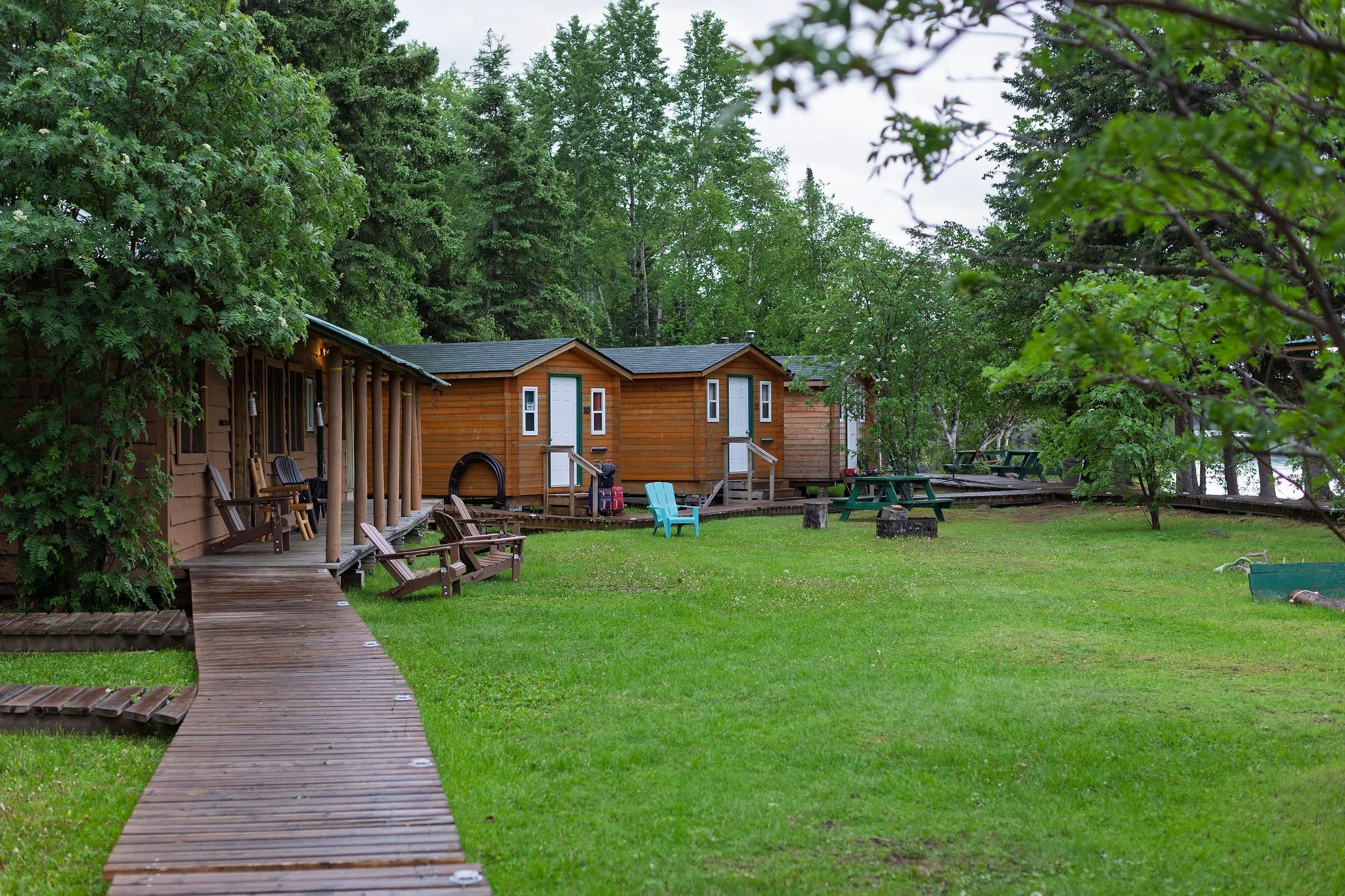 A lakeside scene with small wooden cabins surrounded by tall green trees and a well-maintained grassy area. There are picnic tables and Adirondack chairs on the lawn, with a wooden boardwalk leading to the cabins. The sky is overcast, and the trees a