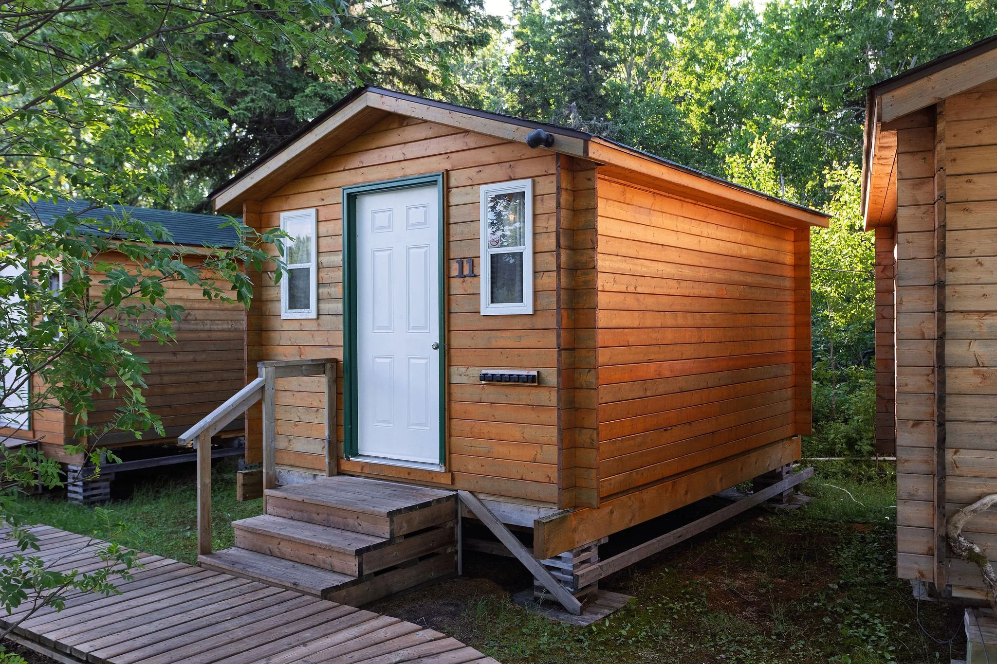 A small private wooden cabin with a white door and two windows, situated in a green wooded area, with steps leading up to the entrance.