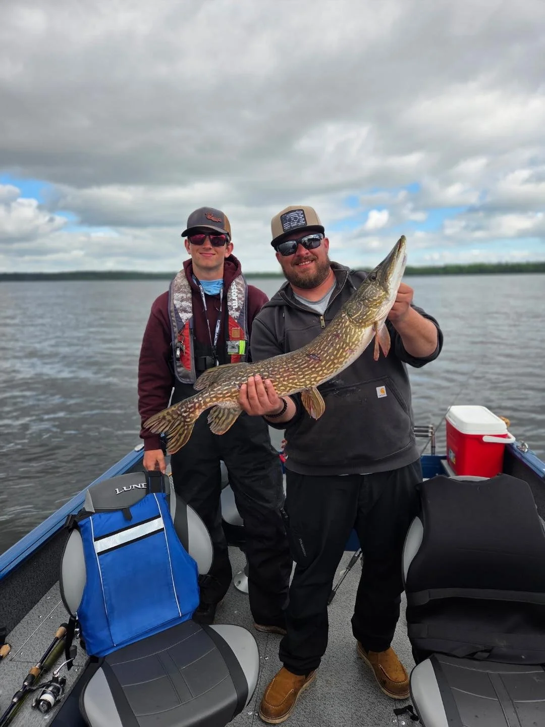 Two men on a boat holding a large fish, with water and cloudy sky in the background.