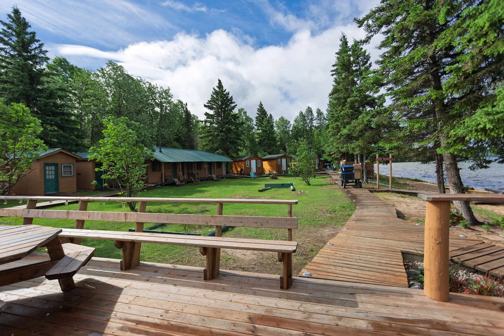 A scenic view of a lakeside area with wooden cabins, a wooden deck with picnic tables, a walking path, tall evergreen trees, and a partly cloudy sky.