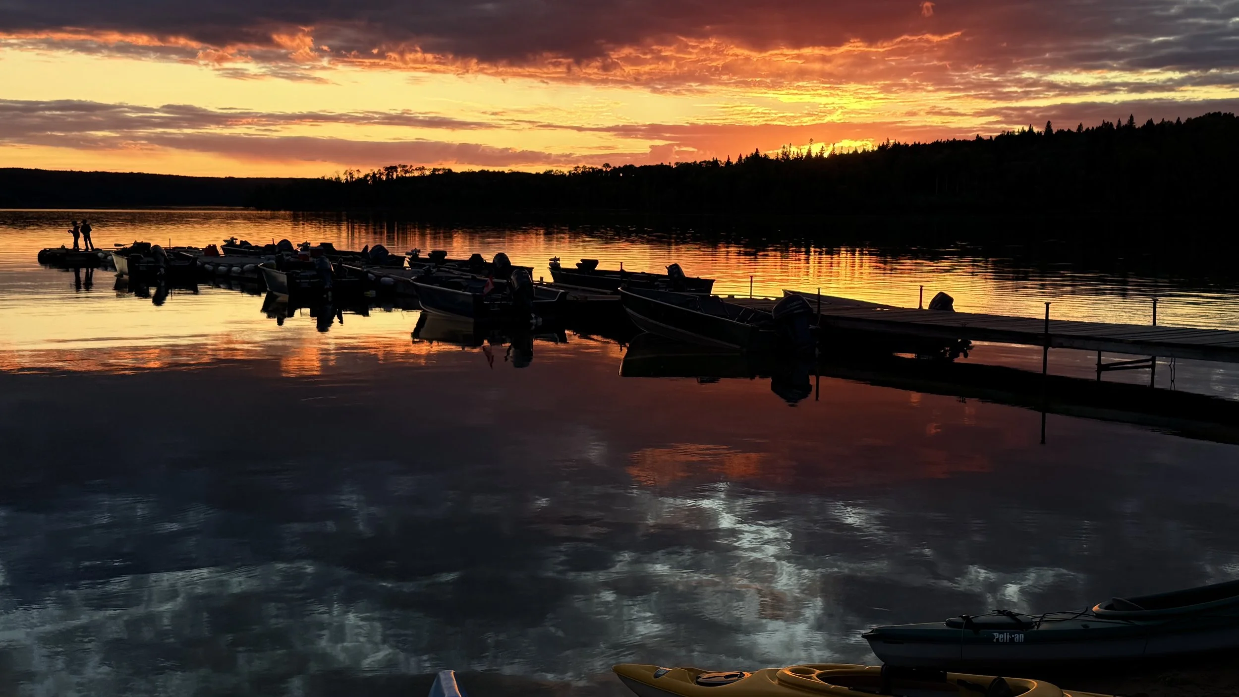 A peaceful lakeside scene at sunset with boats docked along a wooden pier and two people standing on the dock, silhouetted against a colorful sky of orange, pink, and purple with clouds reflected in the calm water.