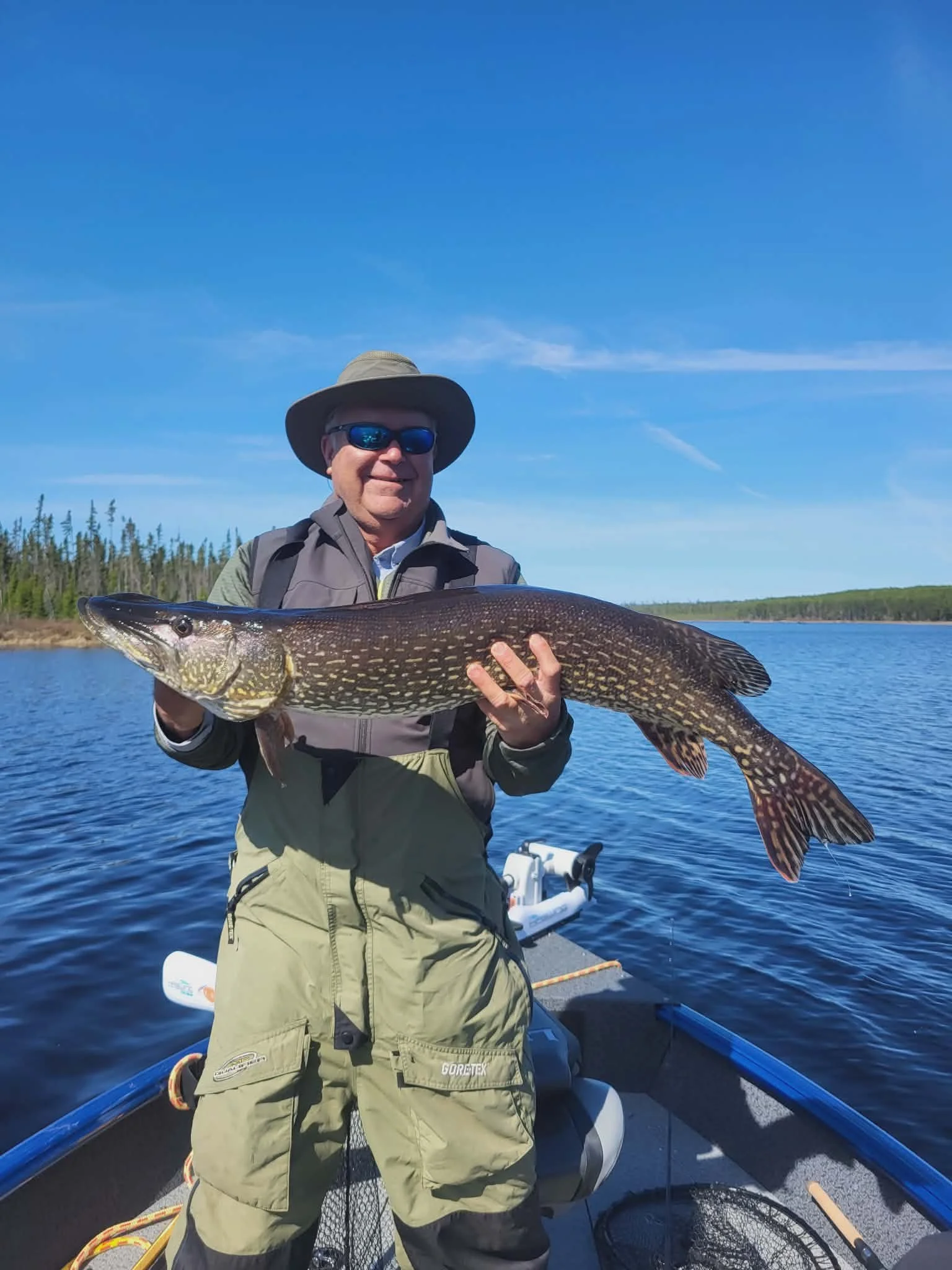 A man in sunglasses, a wide-brimmed hat, a light gray jacket, and green fishing pants is smiling and holding a large fish with a dark, spotted pattern on a boat in a lake, with a forested shoreline and blue sky in the background.