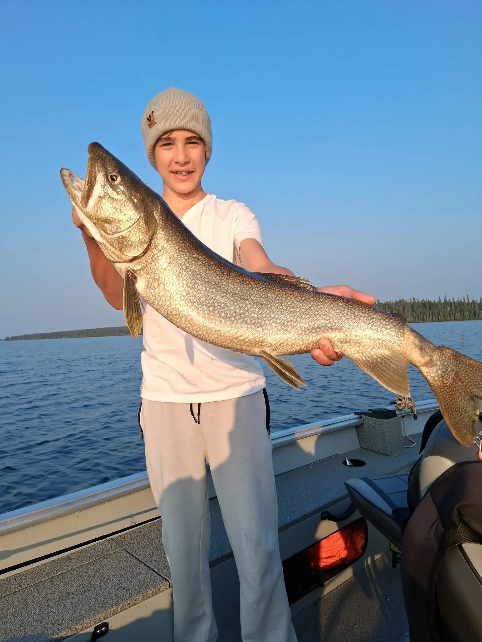 A young boy in a beige beanie and white T-shirt holding a large fish on a boat with water and trees in the background.