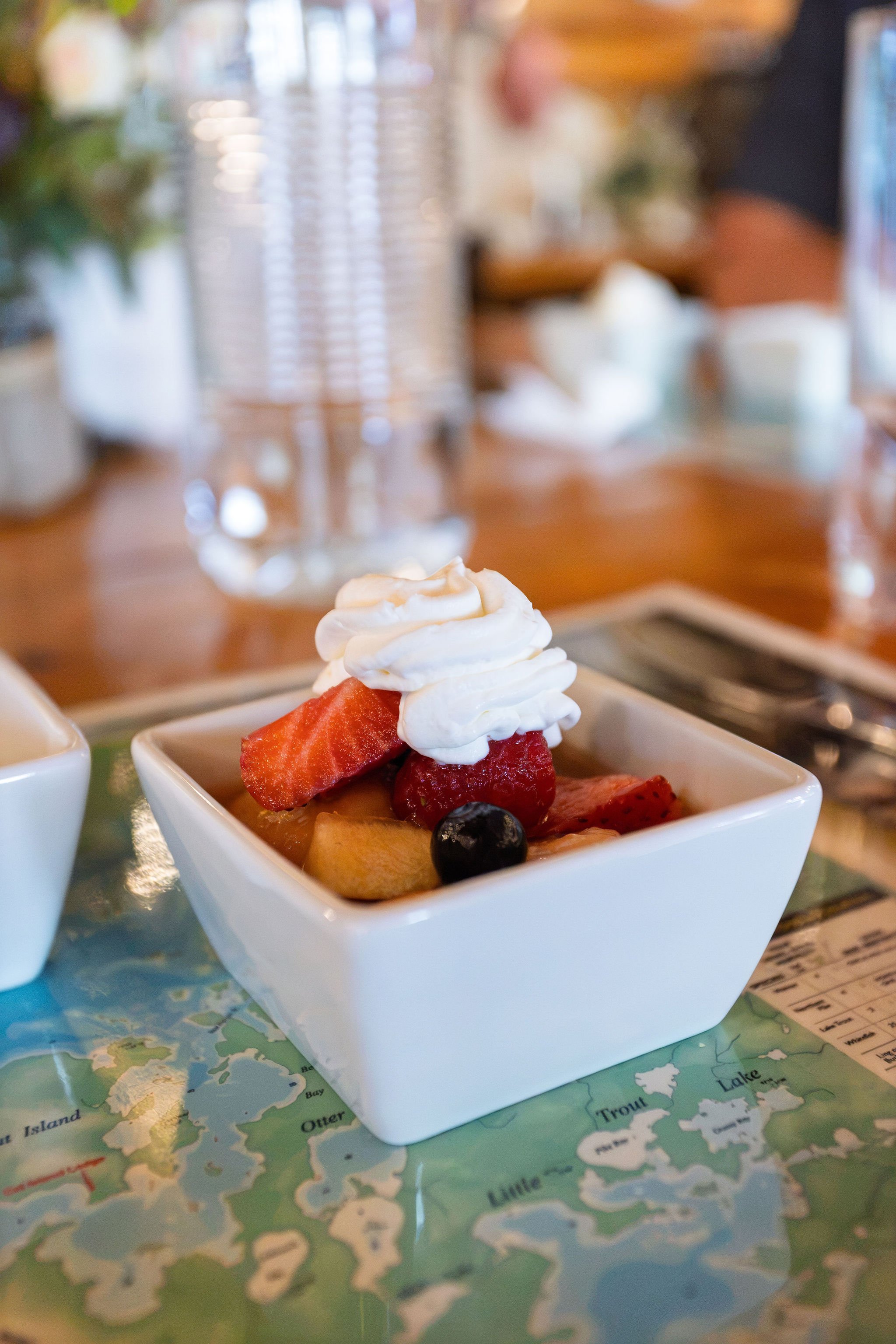 A small white square dish filled with fresh mixed berries and a dollop of whipped cream on top, placed on a map-themed table.