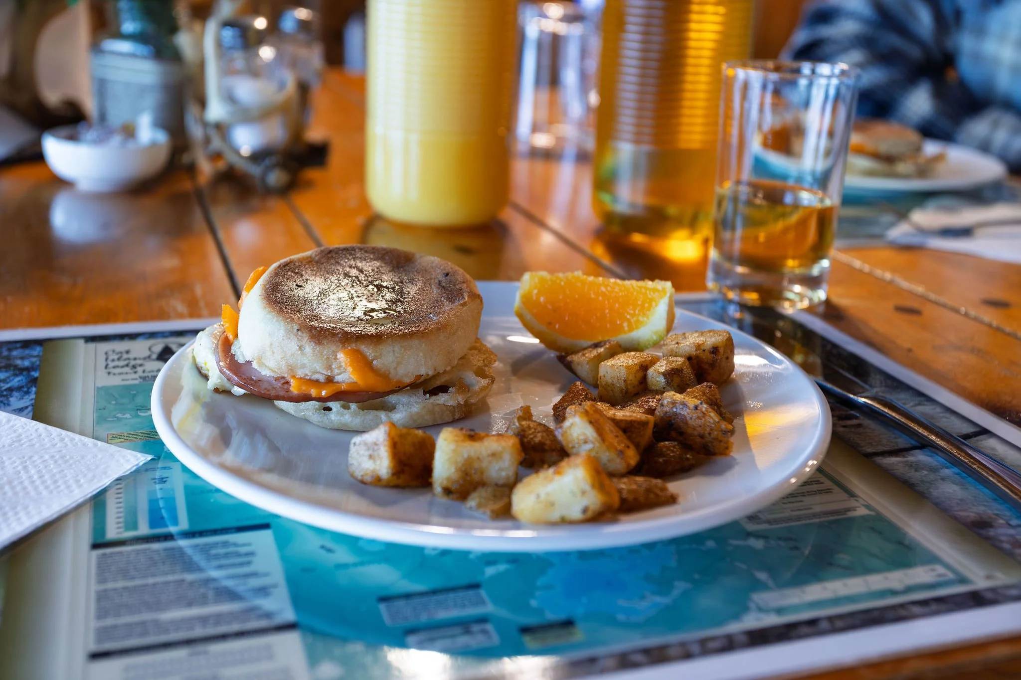 A breakfast plate with an egg muffin sandwich, roasted potatoes, and a slice of orange on a table with drinks and condiments in the background.