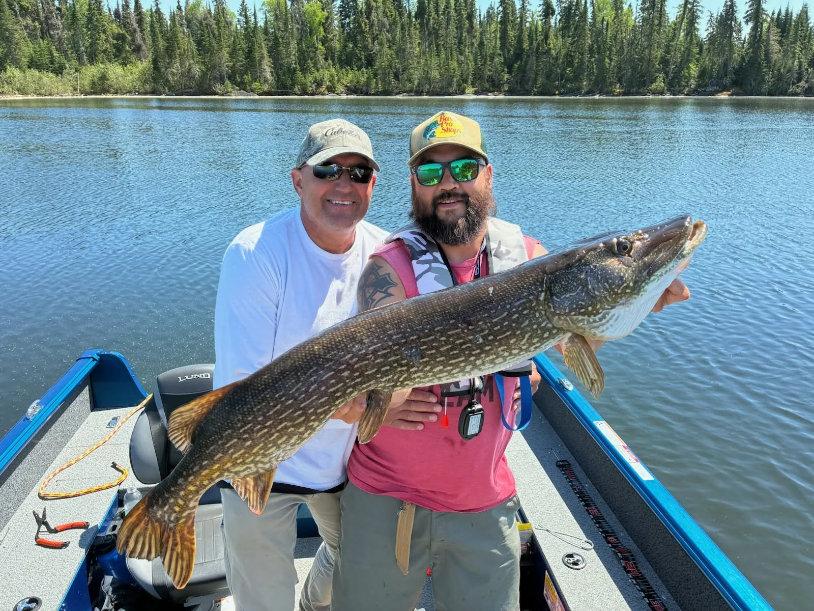 Two men on a boat in a lake, holding a large fish. They are wearing sunglasses and hats, with one man in a pink shirt and the other in a white shirt, surrounded by a scenic view of trees and water.