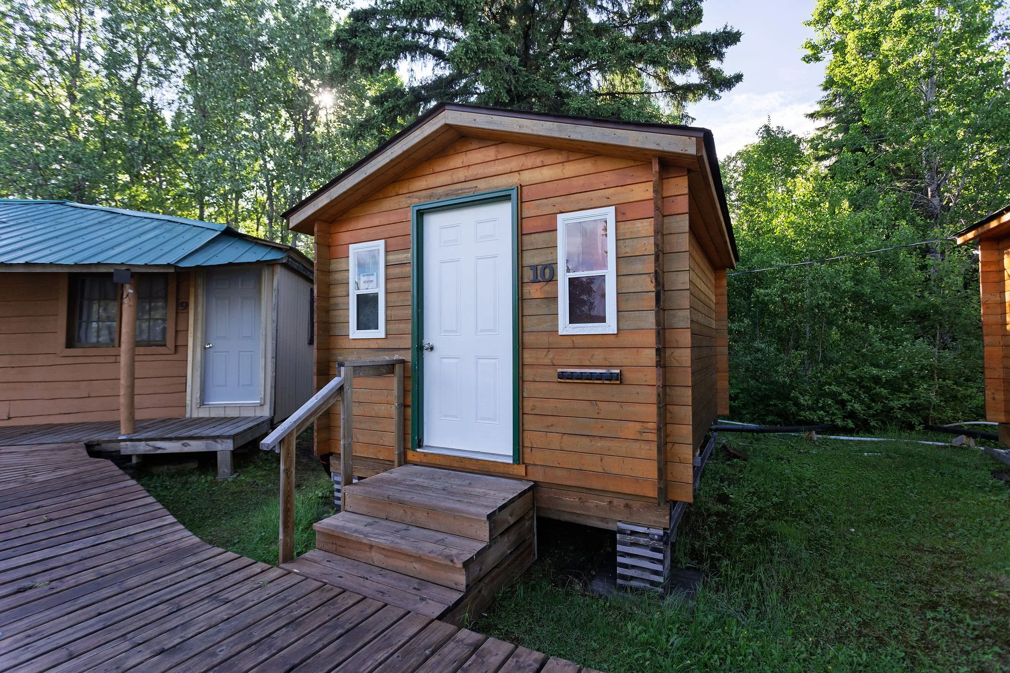 Small wooden house with a white door, two windows, and a small set of stairs, situated in a wooded area with trees and sunlight in the background.