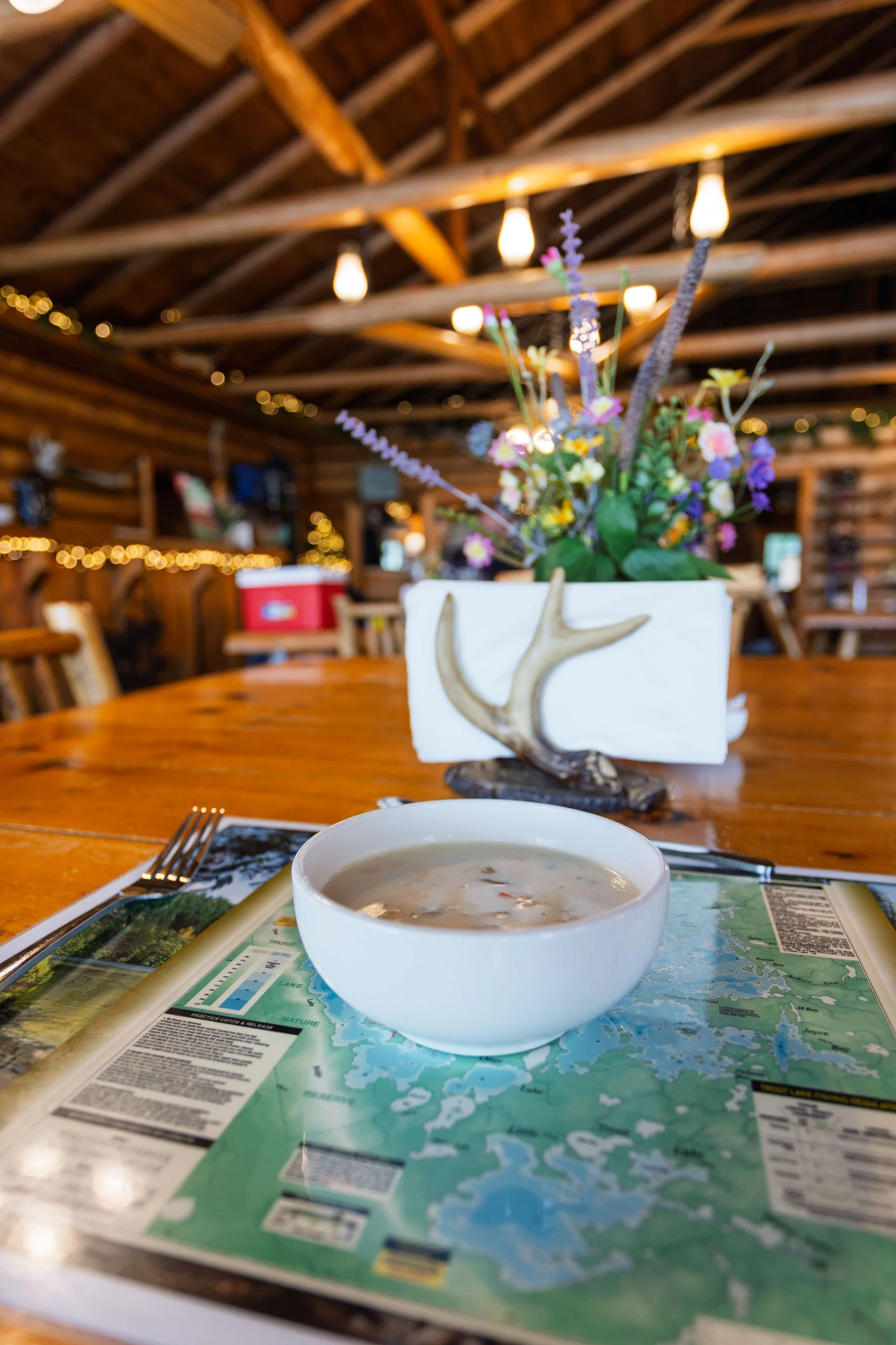 A bowl of soup on an outdoor table, with a map and a fork beside it, and a decorative flower arrangement in the background, under a wooden ceiling with hanging lights.