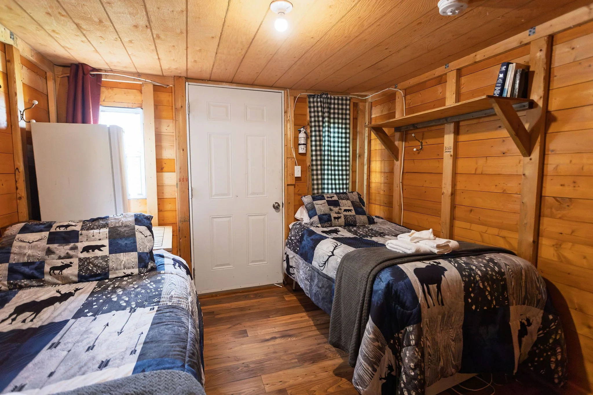 Three beds with black and white outdoor-themed bedding in a rustic wooden cabin room, with a small window, curtains, a shelf with books, and a fire extinguisher on the wall.