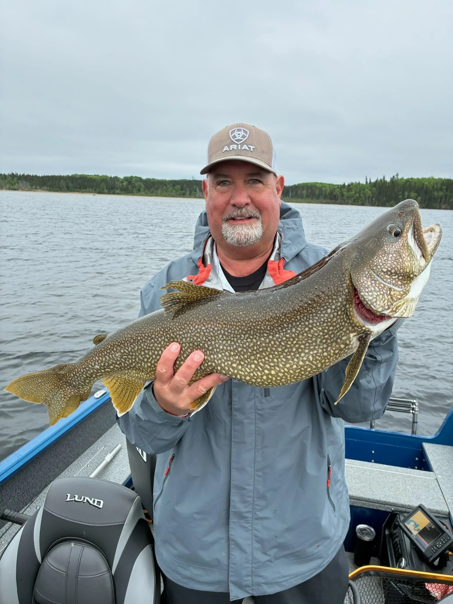 A man holding a large fish on a boat in a lake with a forested shoreline in the background.