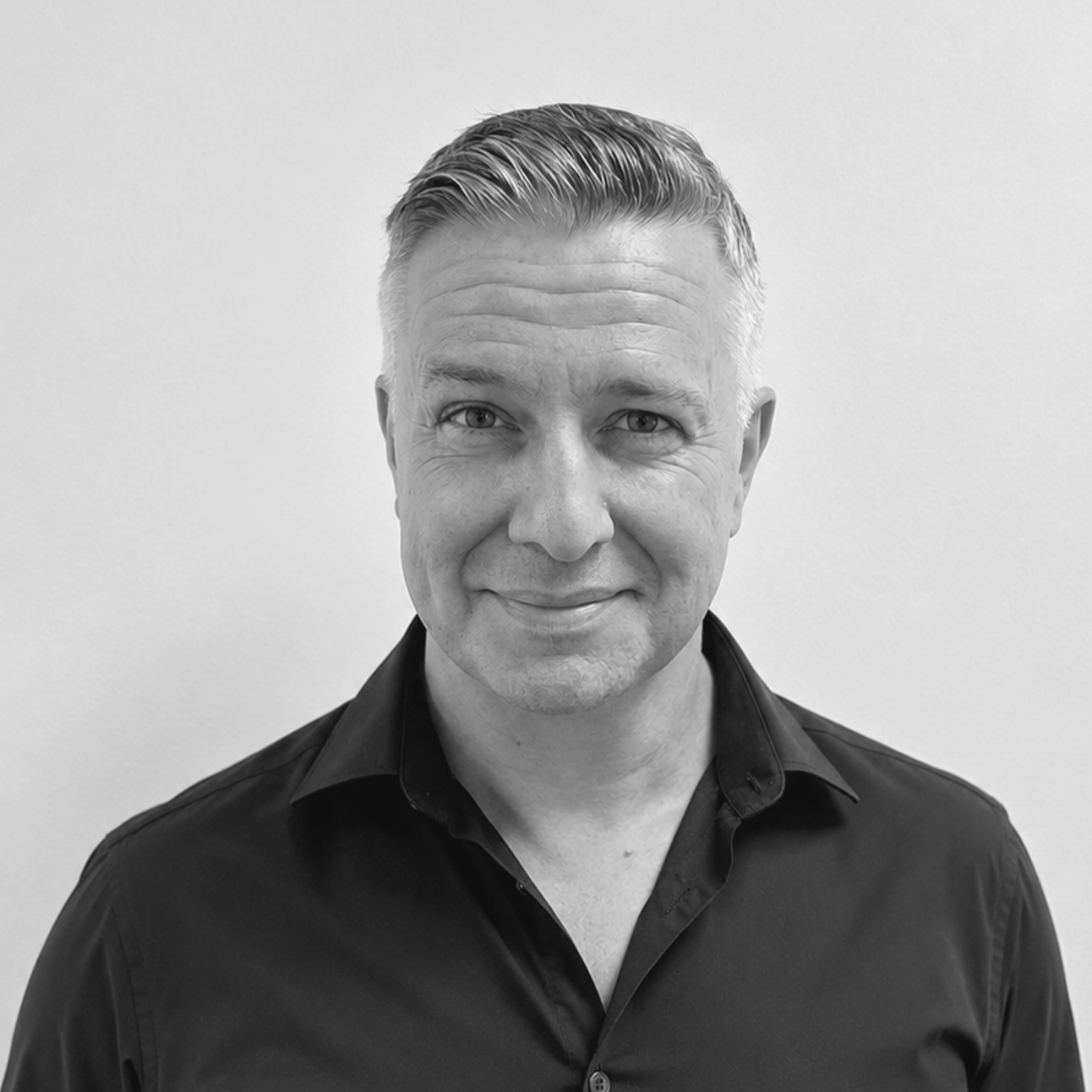 Black and white portrait of a middle-aged man with short, slicked-back hair, wearing a dark collared shirt and a grey blazer, against a plain light background.