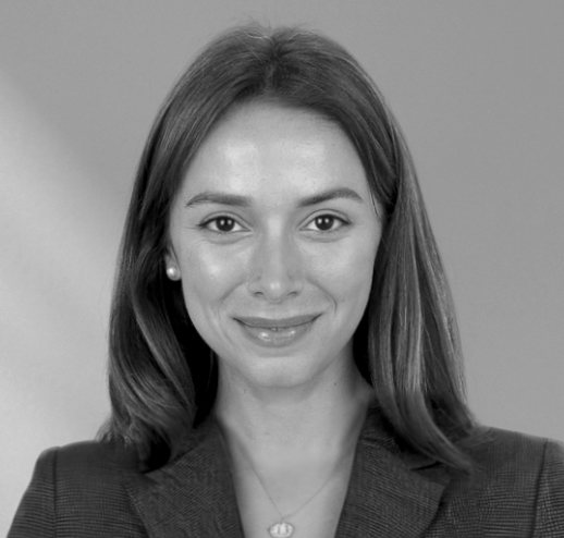 Headshot of a woman with shoulder-length brown hair, wearing a navy blazer and pearl earrings, smiling against a gray background.