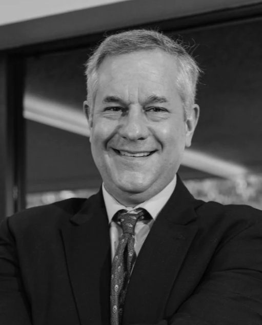 A smiling middle-aged man in a business suit with crossed arms, standing indoors with large windows and green trees visible outside.