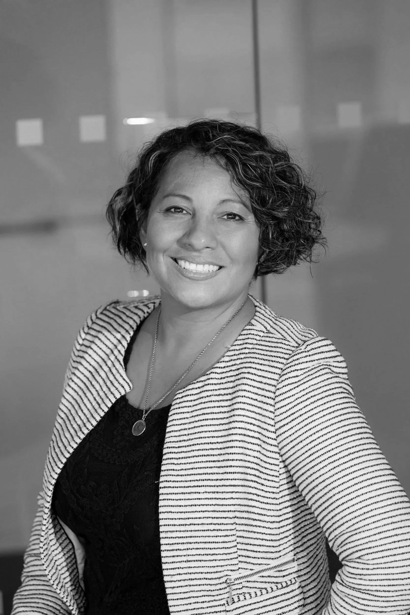 A woman with short, curly brown hair smiling in a professional setting, wearing a black top, a striped blazer, and a gold necklace.