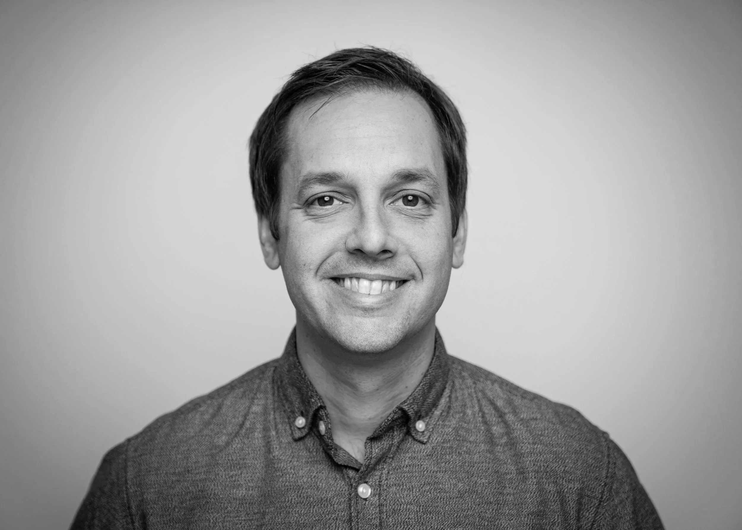 Headshot of a smiling man with brown hair wearing a dark button-up shirt against a plain gray background.