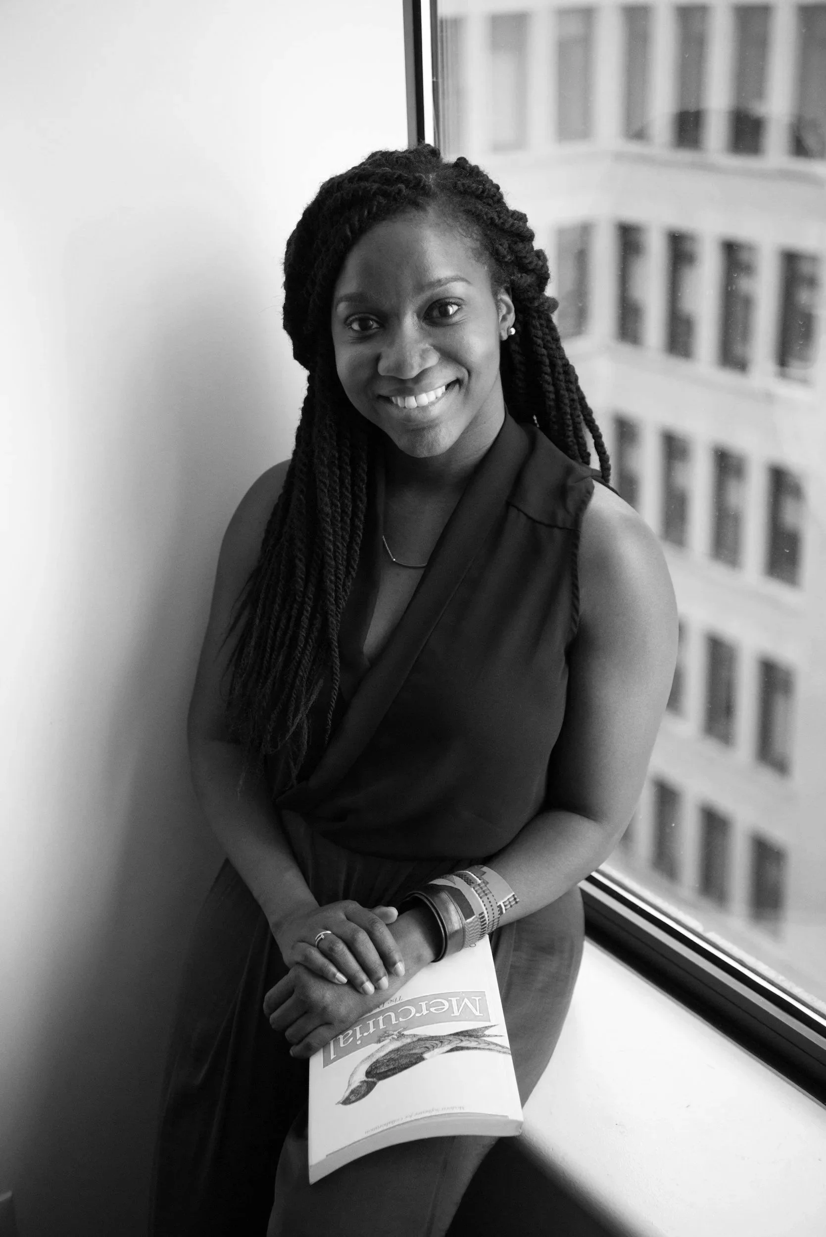 A smiling woman with braided hair wearing a black sleeveless top and pinkish-brown pants, wearing colorful bracelets, holding a magazine titled 'MeCuLia' near a large window with a cityscape background.