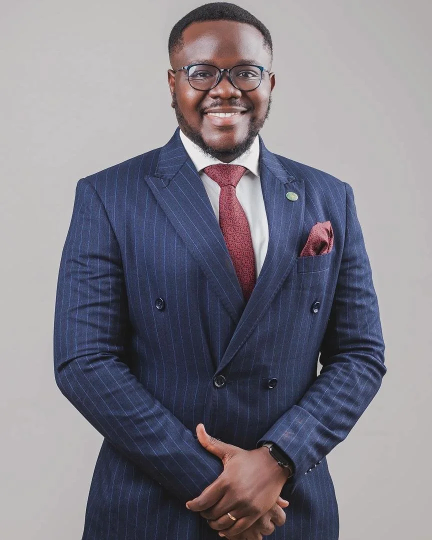 A smiling Black man in a navy pinstripe suit with a red tie and pocket square, wearing glasses, standing against a gray background.