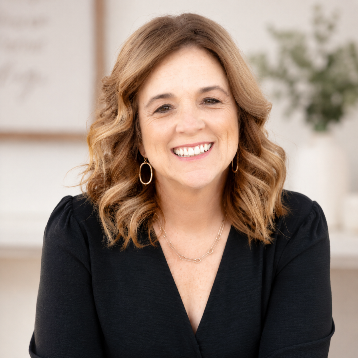 Portrait of a woman with shoulder-length wavy light brown hair, smiling, wearing a black top, gold hoop earrings, and a delicate gold chain necklace, in an indoor setting with blurred background.