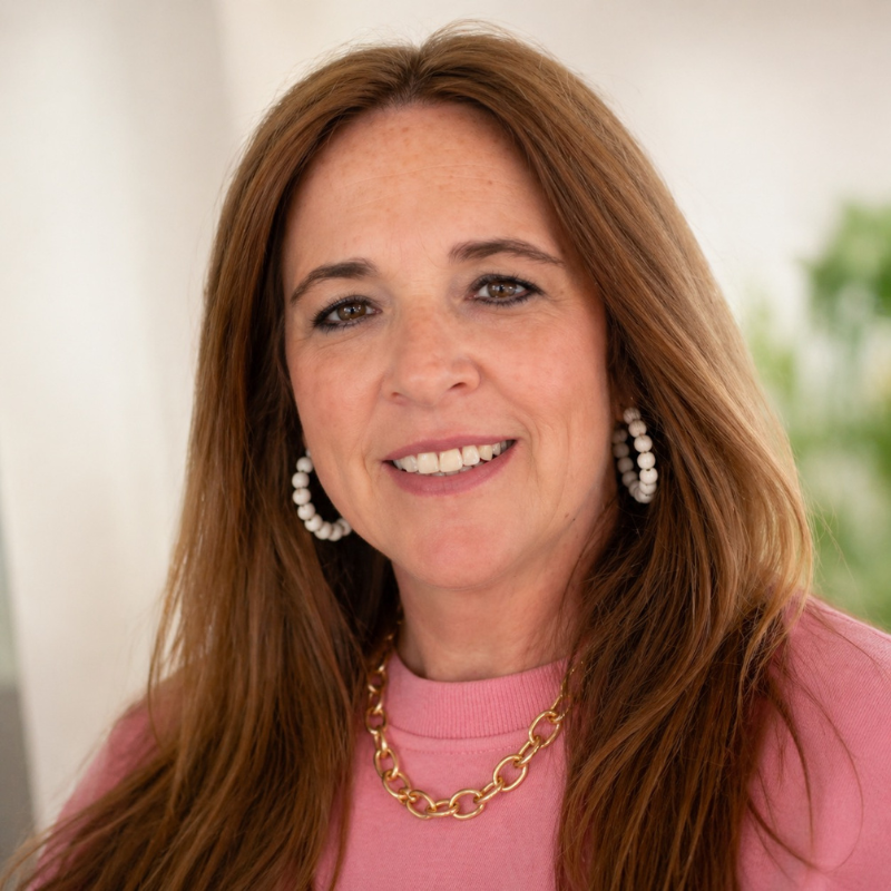 A woman with long, wavy red hair wearing a pink top, white beaded hoop earrings, and a gold chain necklace, smiling outdoors with greenery in the background.