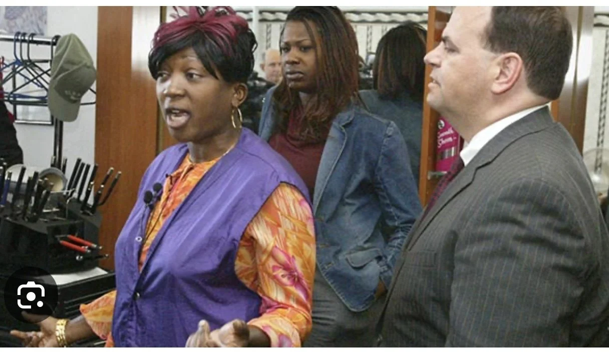 Three people in a store, a woman speaking and gesturing, a man in a suit listening, and a woman with a concerned look in the background.