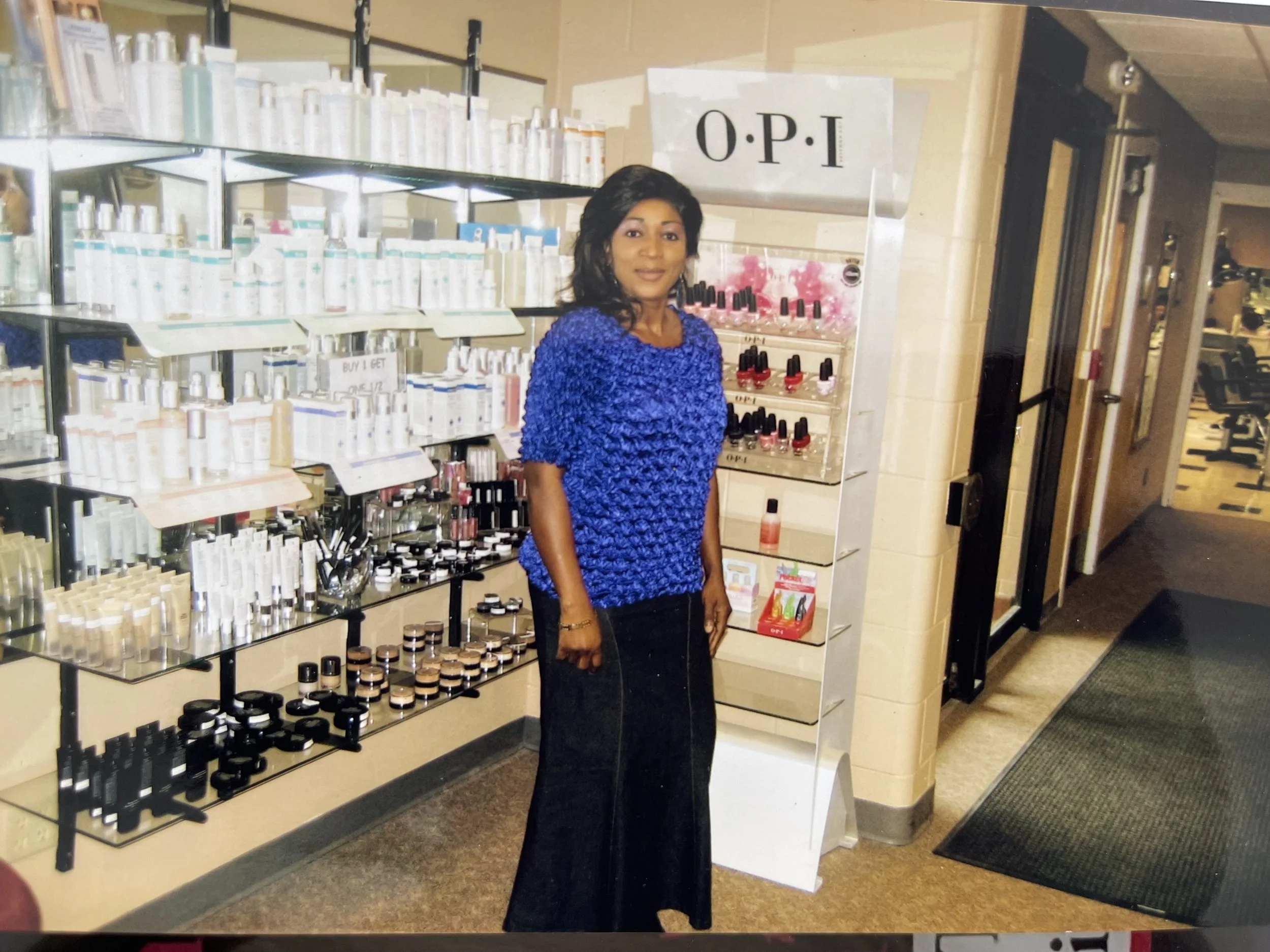 A woman with dark hair wearing a blue textured top and black skirt standing inside a store with makeup products on shelves behind her.