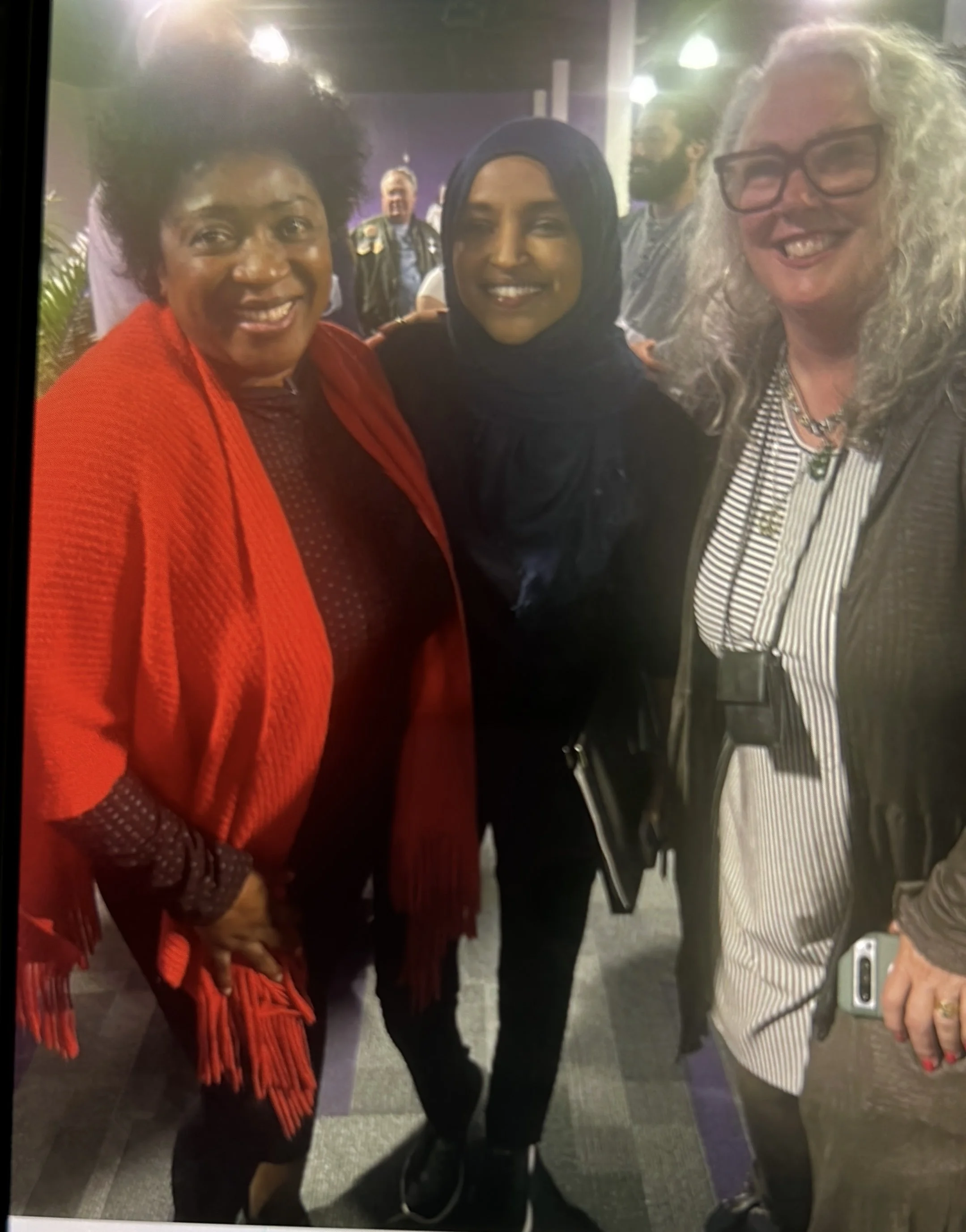 Three smiling women standing close together at an indoor event, with other people in the background.