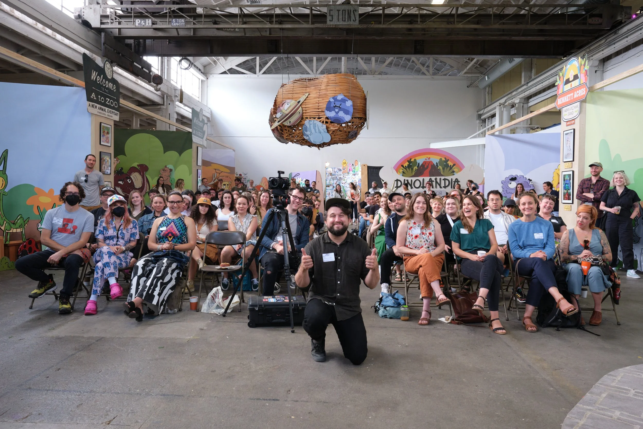 A large group of people seated in chairs inside a spacious venue with industrial ceilings and colorful murals on the walls. A person kneels at the front giving thumbs up, with a camera on a tripod nearby. Various attendees are casually dressed, with 