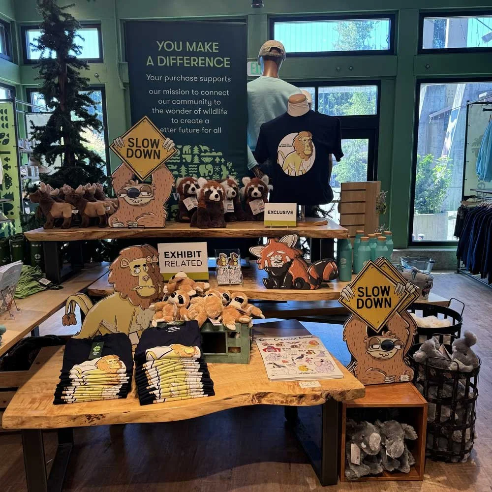 Store display featuring plush animals, wildlife-themed merchandise, T-shirts with animal graphics, and "Slow Down" sloth signs, promoting a conservation mission.