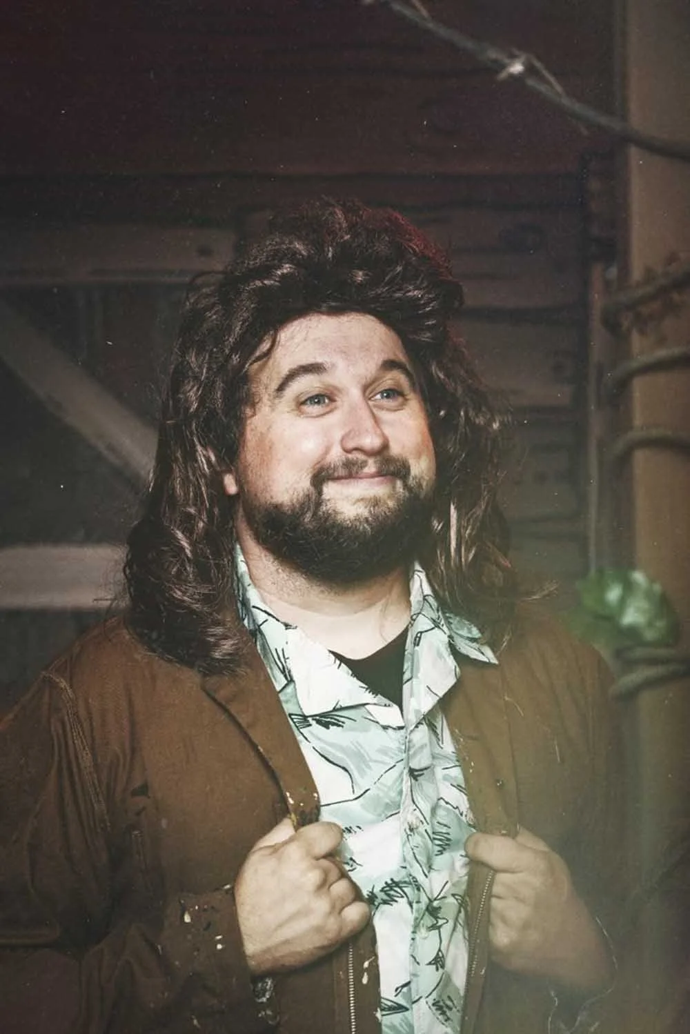 Mike with long hair and a beard smiling, holding the collar of a brown jacket over a patterned shirt.