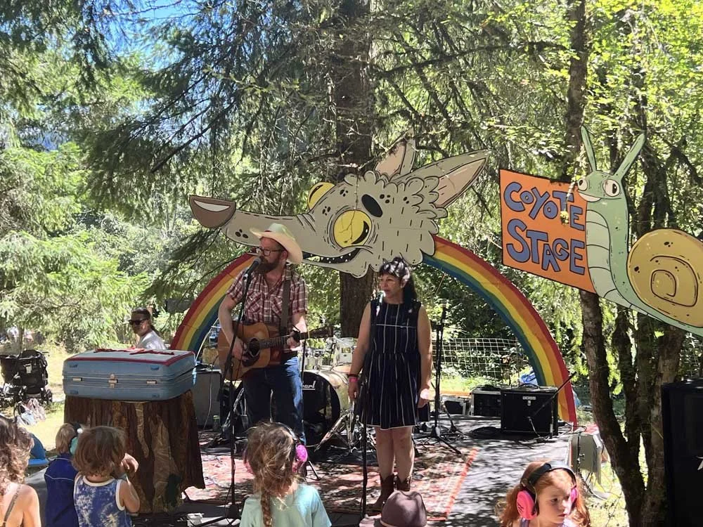 Outdoor musical performance on a small stage decorated with rainbow, coyote, and snail illustrations. A man plays guitar while a woman sings, surrounded by trees. Children with ear protection watch in the audience. A sign reads 'Coyote Stage.'