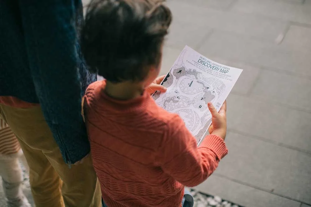 Child holding a discovery map, standing beside an adult, wearing an orange sweater.