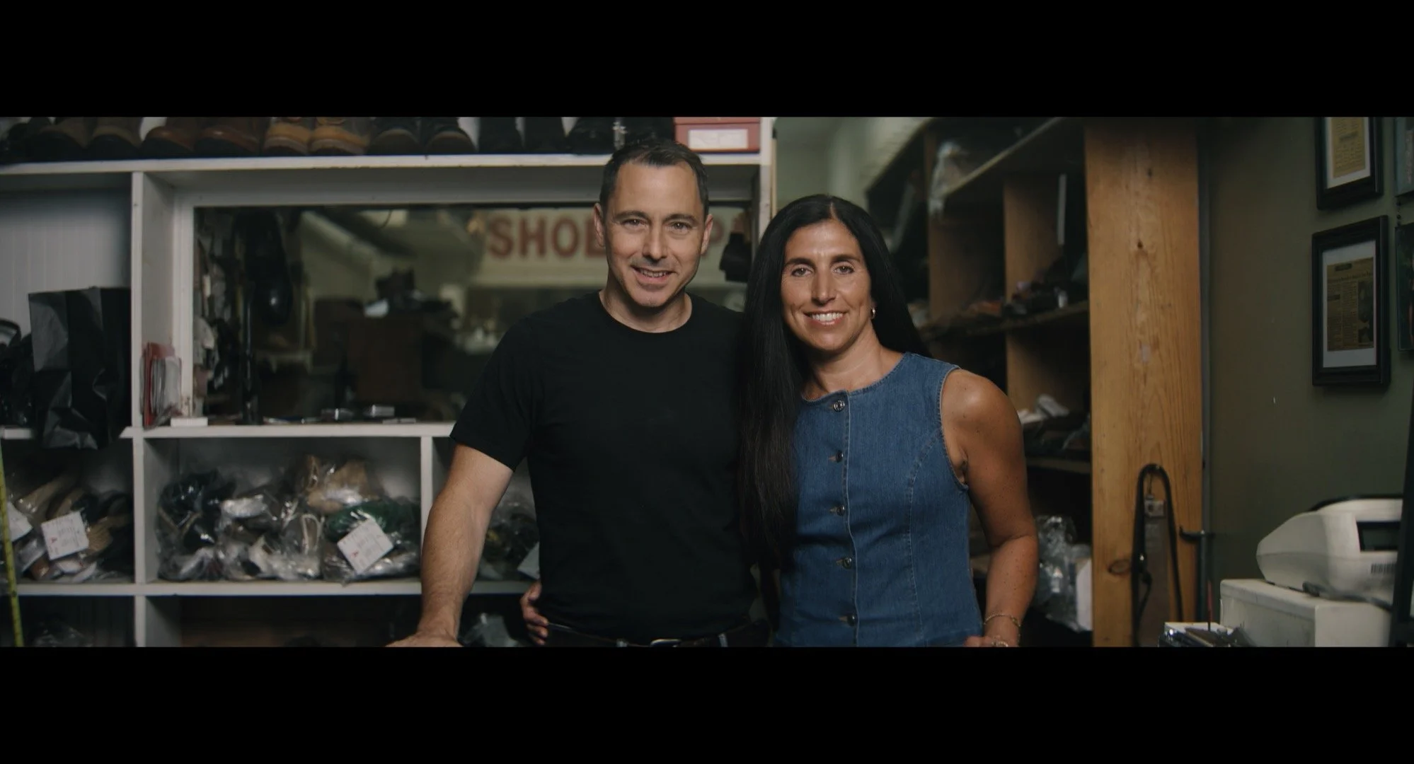 A man and a woman smiling and posing together inside a shoe store, with shelves of shoes and framed pictures in the background.