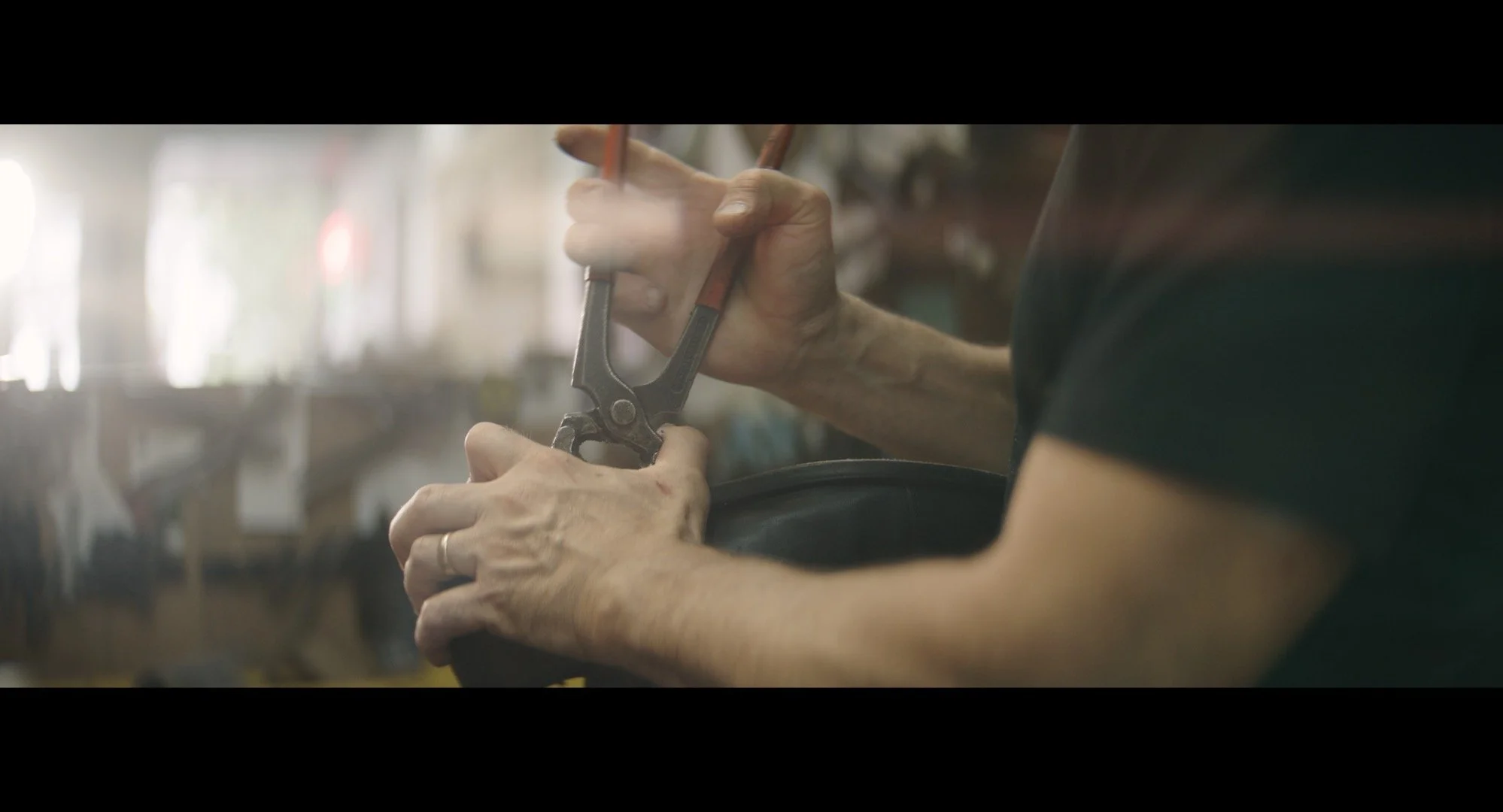 Close-up of elderly person's hands fixing a pair of pliers. The background shows a blurred workshop or garage setting.
