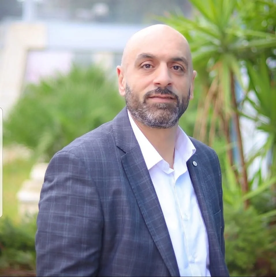 A man with a beard wearing a dark plaid blazer and a white shirt stands outdoors with green plants in the background.