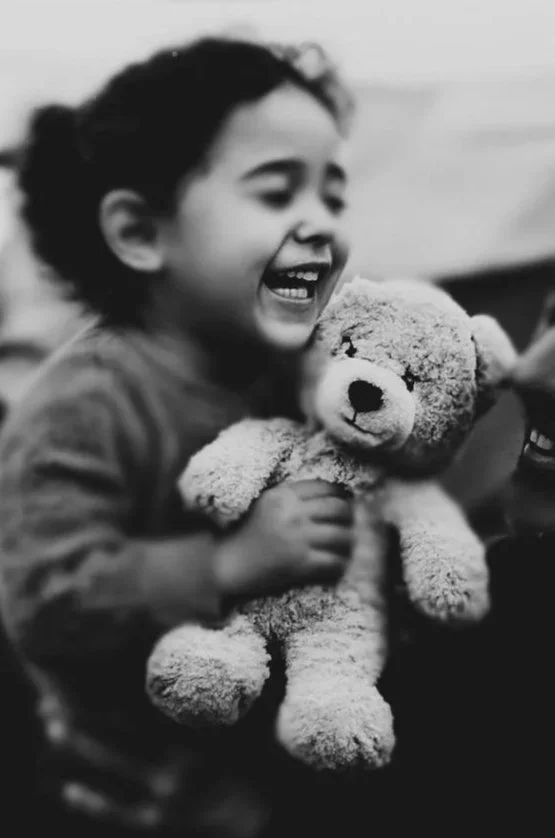 Santa Claus with a white beard and red suit, sitting at a wooden cart with the words 'Give Gaza' written on it, surrounded by toy bottles and a teddy bear, during sunset in a city with minarets and burning buildings, with two doves flying in the sky.