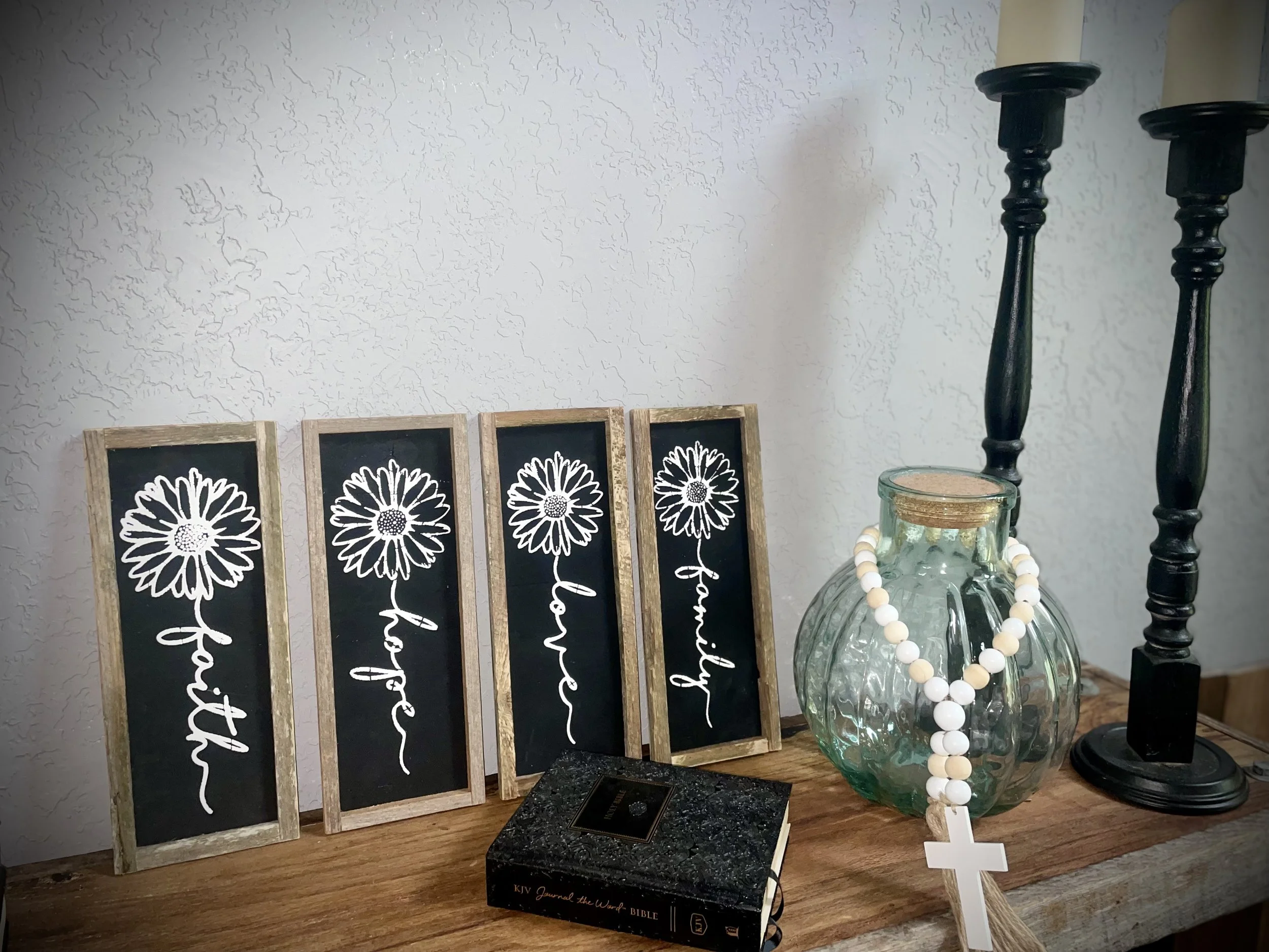 Decorative display on a wooden table with four framed wooden signs with white daisies and words 'faith,' 'hope,' 'love,' and 'family,' a glass vase with a beaded rosary, a black Bible, and two black candlesticks.