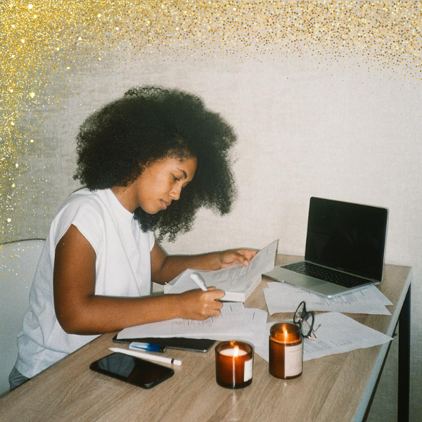 A young woman with curly hair sitting at a desk, studying with papers, a laptop, and a pen, surrounded by candles and eyeglasses.