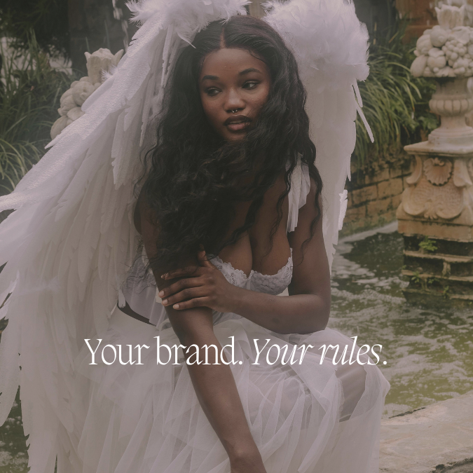 A woman with long curly hair dressed as an angel in a white dress with large white feathered wings, sitting by a pond with a stone fountain in the background, with the text 'Your brand. Your rules.'