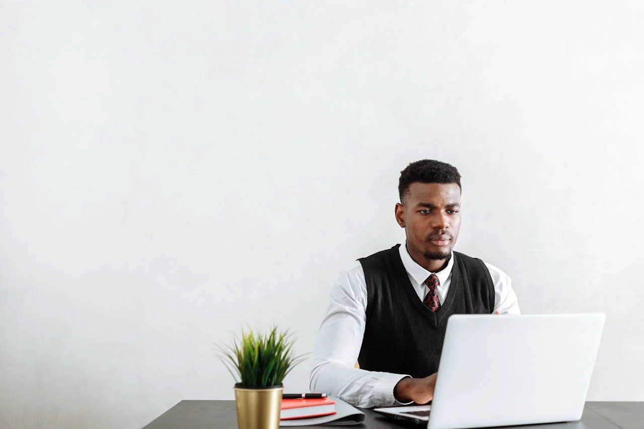 Young man in business attire working at a desk with a laptop, a notebook, a pen, and a potted plant against a plain white wall background. Blueprint studio creative artist web design